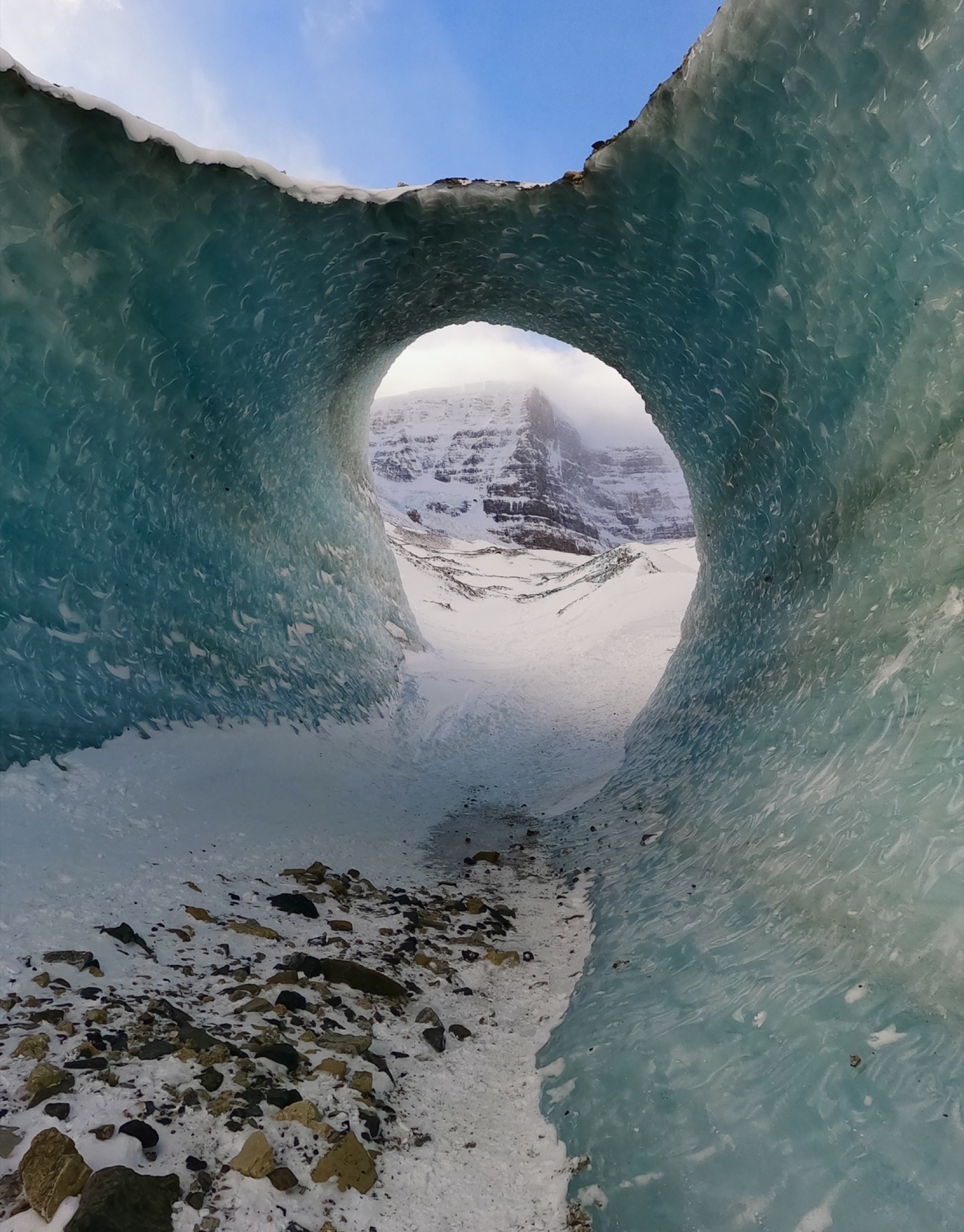 Looking through a teal glacier ice tunnel toward a snowy valley, Banff